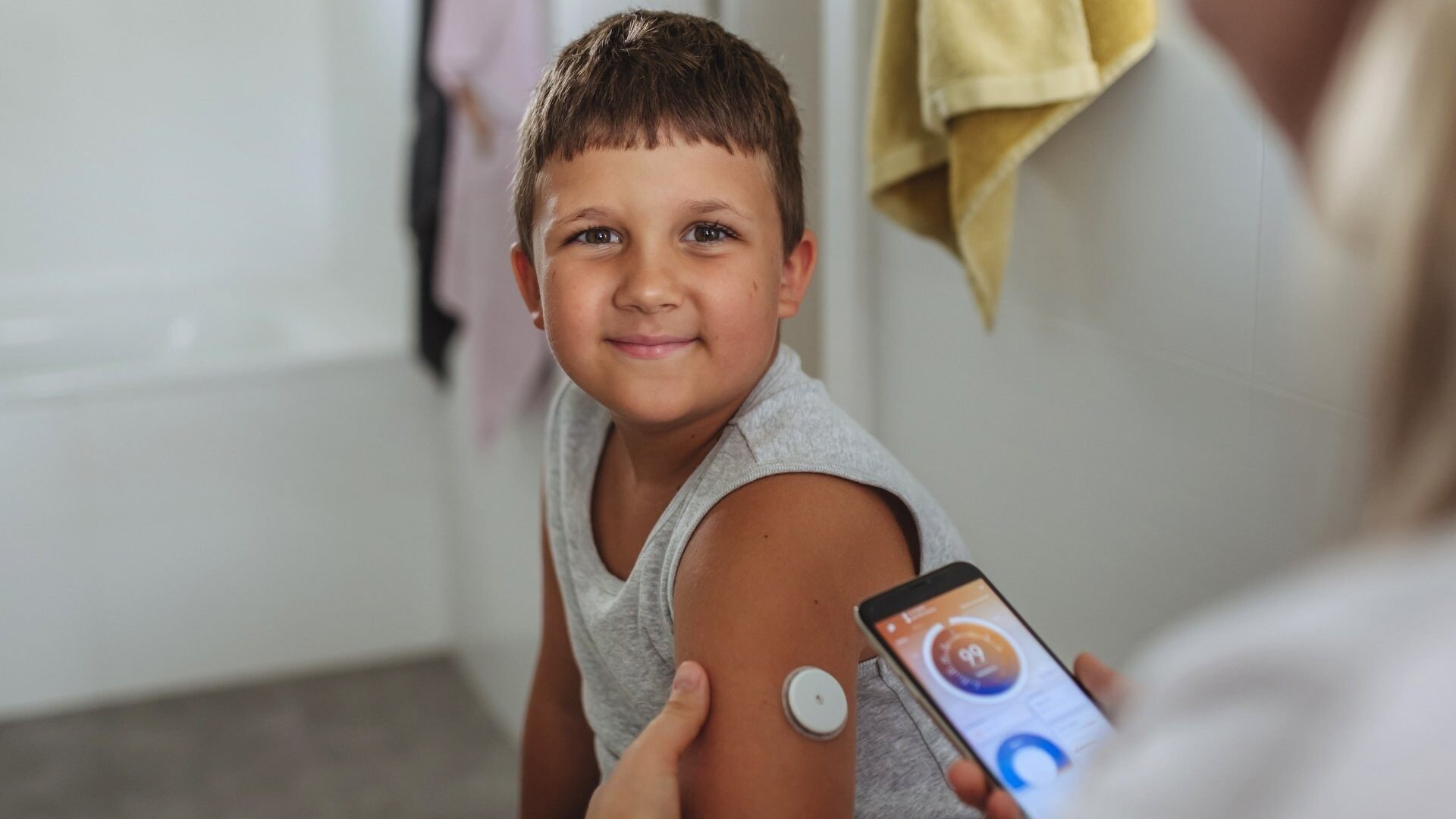 Boy-smiling-at-camera-with-cgm-mother-checking-glucose-with-smartphone
